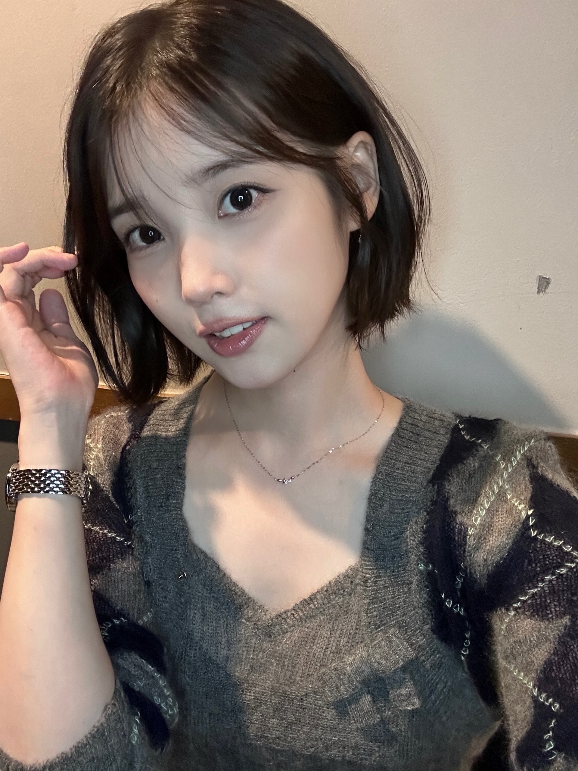 단발머리의 인물이 회색 니트를 입고 머리카락 옆에 손을 올린 근접 사진